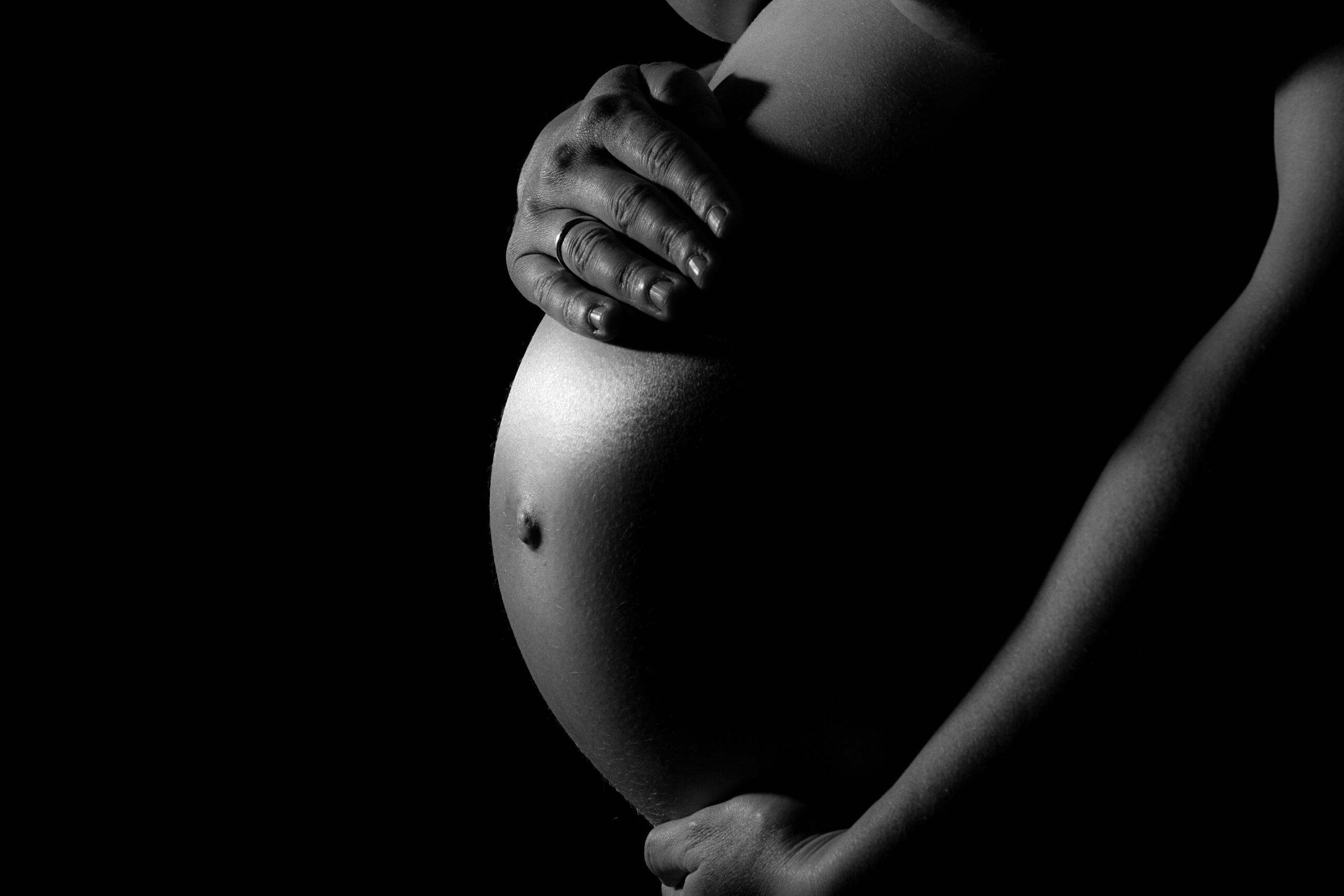 Mittelteil einer schwangeren Frau, die ihren Bauch vor einem schwarzen Hintergrund berührt